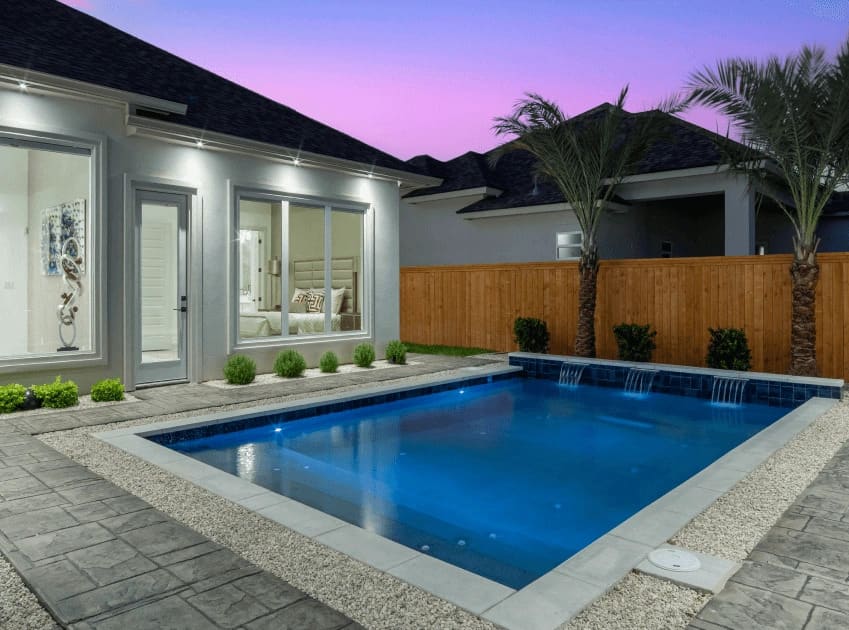 Modern backyard featuring a rectangular swimming pool, surrounded by stone patio, lush greenery, and a wooden fence at dusk.