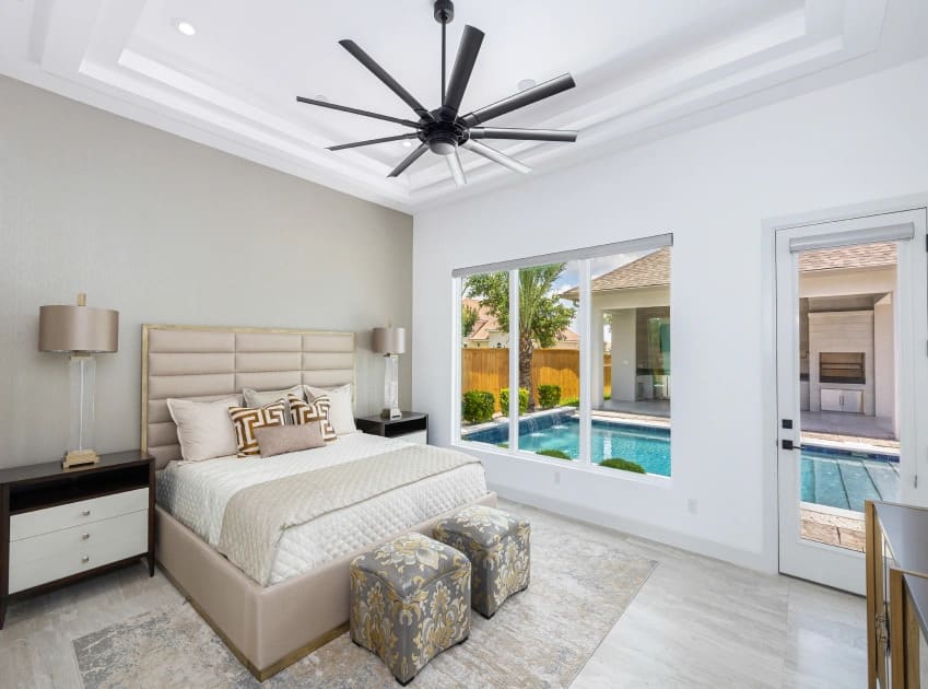 Modern bedroom featuring a large bed, decorative stools, and a view of a pool through a large window.