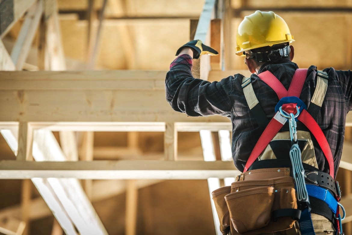 Wood Construction Worker. Caucasian Builder Wearing Safety Harness and the Wooden House Structure. Industrial Theme.