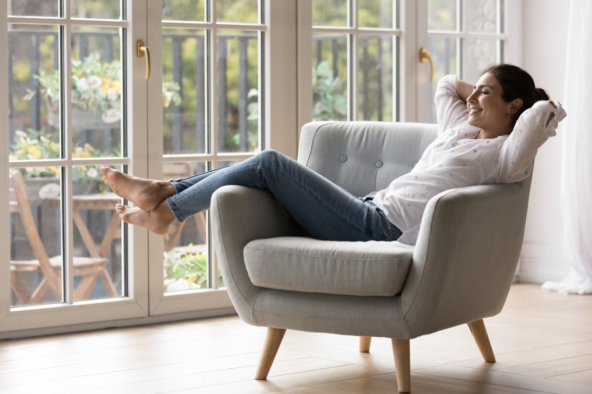 Pretty Indian woman chilling resting on comfortable armchair near panoramic windows in light sunny modern living room, female closing eyes enjoy free time and fresh conditioned air inside cozy house