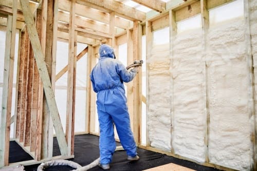 Worker spraying polyurethane foam insulation into timber wall cavities inside unfinished house. Interior construction scene, thermal sealing, weatherization and energy efficient building concept