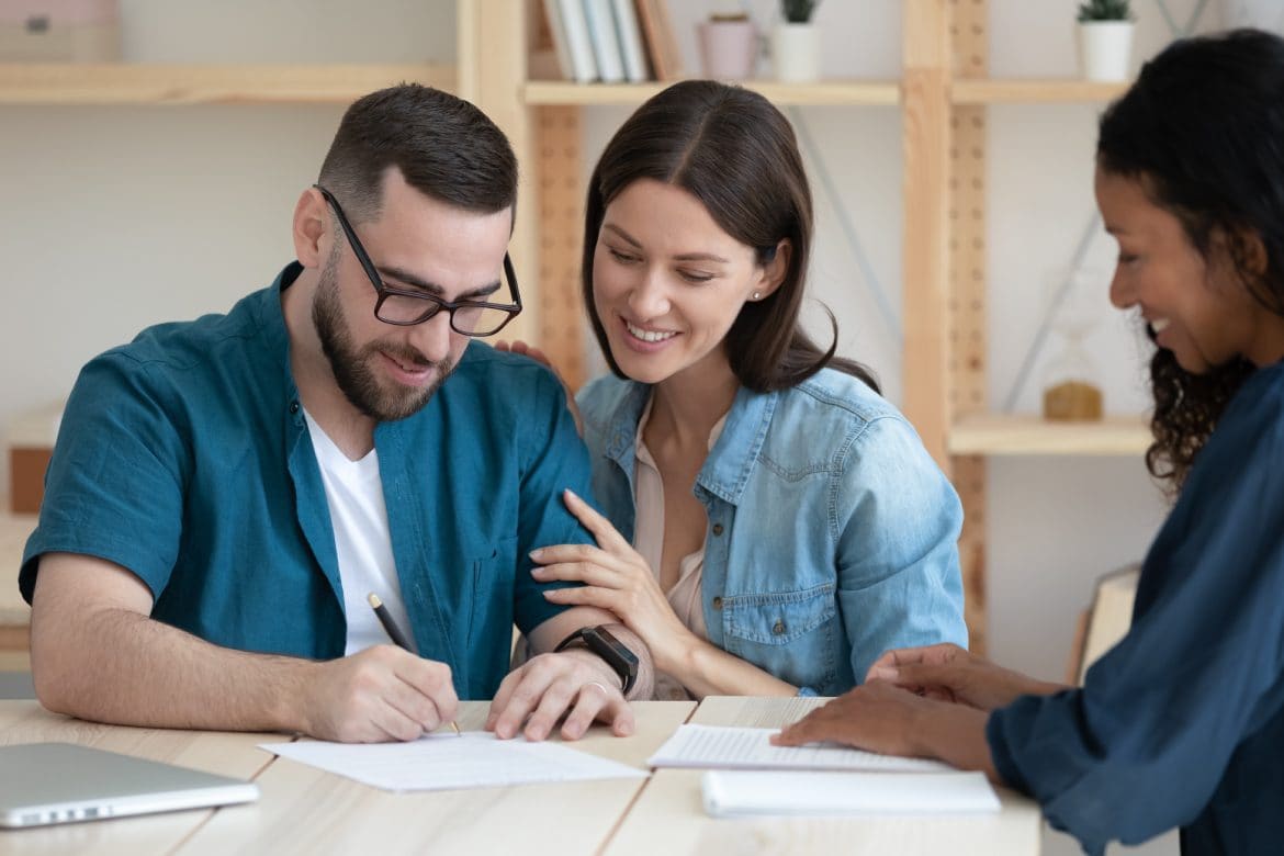 Smiling young family clients make purchase deal sign insurance contract at meeting with african american estate agent. Happy millennial couple reading document about buying house on loan.
