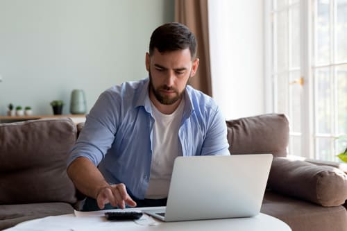Serious millennial man sit on sofa do paperwork, pay taxes and household utility bills on-line using e-bank app on laptop, manage budget, planning savings, investments looks focused. Finances