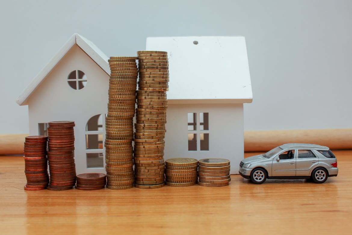 A conceptual image featuring various Euro coins piled in front of small wooden toy houses and a miniature car. This composition represents the significant financial effort, long-term saving strategy, and investment required to achieve the dream of home ownership and personal transport in the current economy.