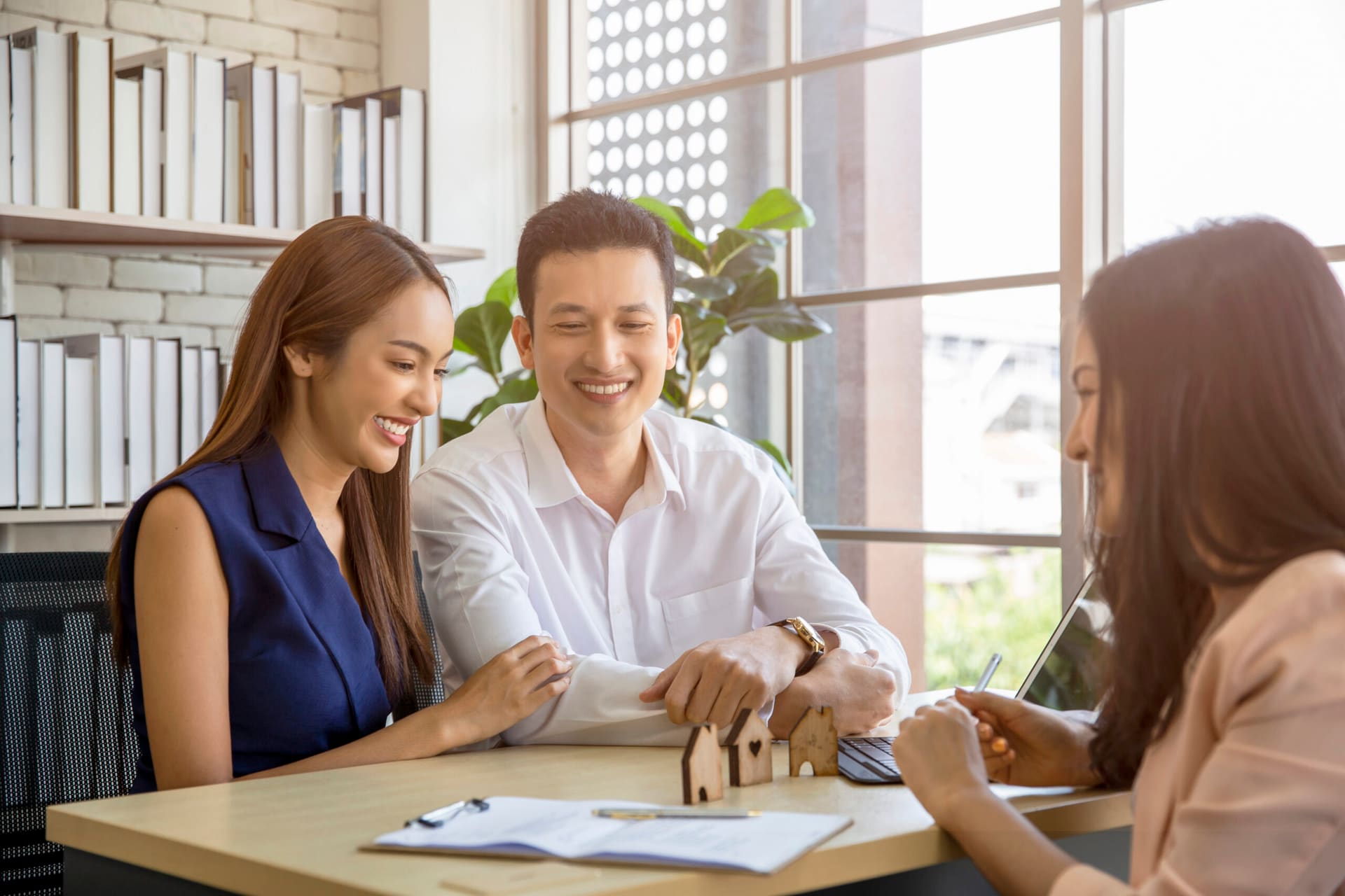 A couple smiles while discussing plans with a real estate agent at a desk, with a model house and paperwork in view.